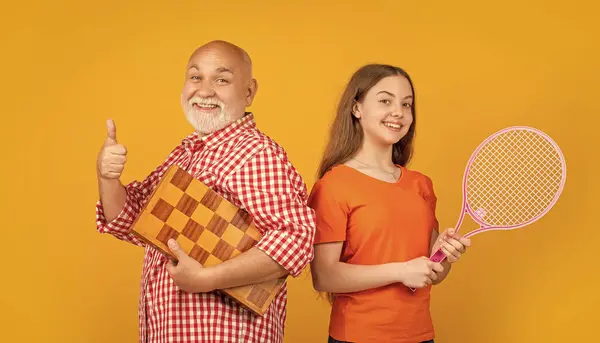 stock image cheerful girl with grandfather with badminton racket and chess on yellow background.