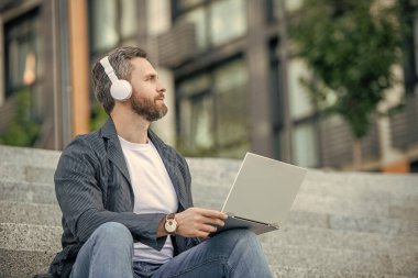 Man Webinar açık havada oturuyor. Adam sokakta internetten internete düştü. Man Webinar dışarıda çevrimiçi. İnternette kulaklıklı bir adamın fotoğrafı..