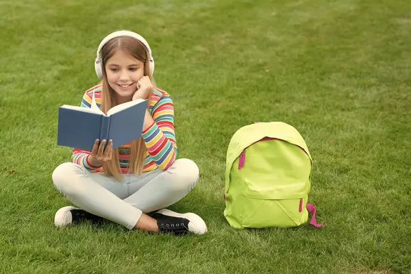 Happy girl learning English language listening to audio course in headphones sitting on grass outdoors, school.
