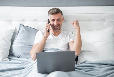 Başarılı bir yetişkin telefonda konuşuyor ve laptopla yatakta çalışıyor..