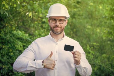 Man foreman in hardhat giving thumbs up showing blank contact card outdoors, copy space.