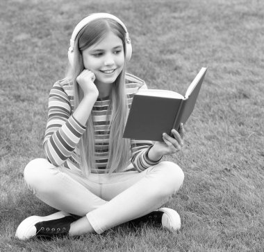 Kulaklıklı mutlu genç kız yeşil çimlerin üzerinde oturmuş kitap okuyor, okul....