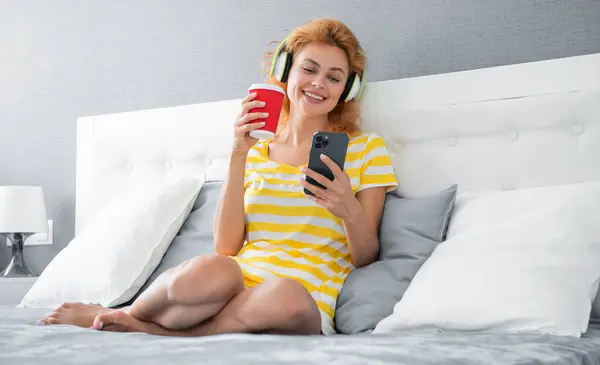 Happy woman reading news on mobile phone at home, mobile phone. Relaxing woman listening to music on mobile phone while drinking tea on bed.