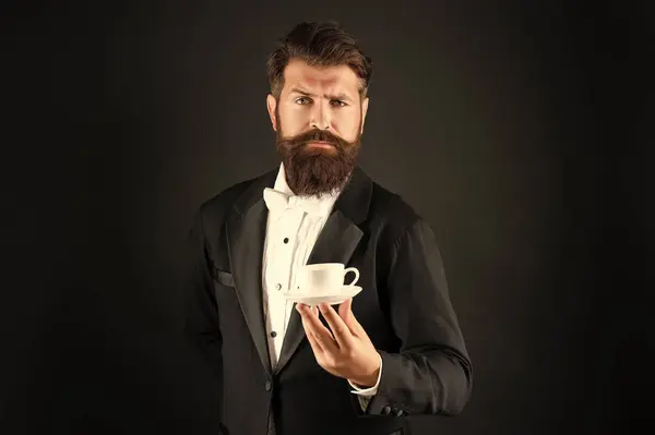 mature man in tuxedo bow tie with coffee cup. waiter in formalwear serving coffee on black background.
