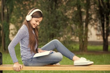 concept of audiobook. Happy teenage girl reading and listening to audiobook in headphones after school outdoors, audiobook.