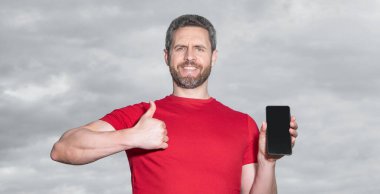 man showing phone app on sky background, thumb up. man showing phone app outdoor. man showing phone app wear red tshirt. photo of man showing phone app.