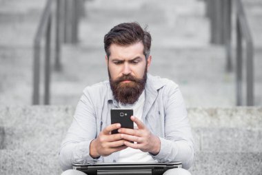 Serious guy chatting on mobile phone while sitting on steps outdoors. Chatting online on mobile device. Chatting using mobile apps in smartphone. Mobile communication.