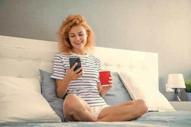 Kadın telefonda internette sörf yaparken gülümsüyor. Evde telefonuyla internette sörf yapan bir kadın. Kadının evinde internet sörfü.