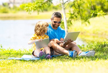 Baba ve oğlunun açık havada online eğitimi var. İnternette öğretmenlik yapan ve parktaki tablet üzerinde çalışan bir öğrenci. Baba ve oğul ders çalışıyor. Oğlu ve babası çocukluk eğitimi almış. Açık hava mesafe öğrenimi.