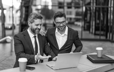Neşeli iş adamları bilgisayarla çevrimiçi ticaret yapıyor. İnternet üzerinden ticaret yapan iş adamlarının fotoğrafı. İki işadamı çevrimiçi ticaret yapıyor. İş adamları açık havada ticaret yapıyor.