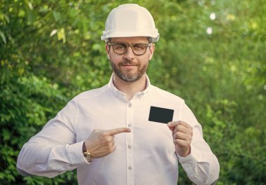 Businessman man in hardhat pointing finger at blank business card outdoors, copy space.