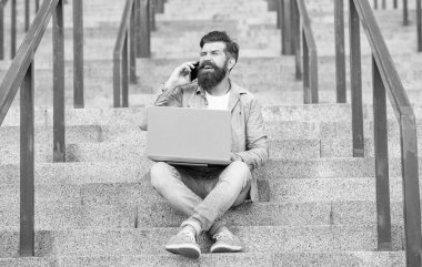 Happy guy talking on mobile phone working on laptop. Smiling guy making mobile call sitting on stairs outdoors. Mobile communication. Mobile calling. Phone conversation.