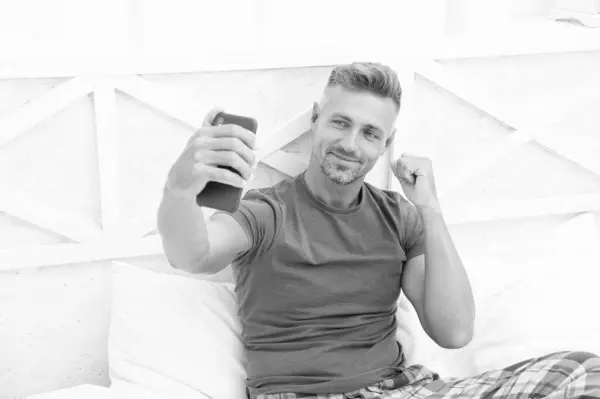 Stock image smiling blogging man with phone in bedroom. blogging man with phone wear shirt. blogging man with phone in bed. photo of blogging man with phone at home.