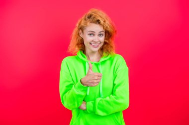 Havalı kapüşonlu giyen kızıl saçlı kız. Pembe üzerine izole edilmiş şık bir kadın. Hareketli moda kız kapüşonlu giyer. Moda kıyafetleri ve tarzı. Kazaklı beyaz bir kadın baş parmağını gösteriyor. Günlük yaşam tarzı.