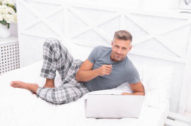 Evde, yatak odasında serbest çalışan neşeli bir adam. Evde dizüstü bilgisayarlı serbest çalışan bir adamın fotoğrafı. Evde serbest çalışan bir adam var. Adam evde, yatakta serbest çalışıyor..