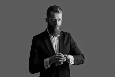 Saati ve smokini kontrol eden dakik bir adam. Smokin modası. Smokinli dakik sakallı adam gri arka planda izole edilmiş saati kontrol ediyor. Smokin giyerken çekilen dakik adam stüdyosu kontrol saati.