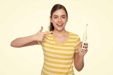 Neşeli genç kadın beyaz arka planda izole edilmiş elektrikli diş fırçasını işaret ediyor..