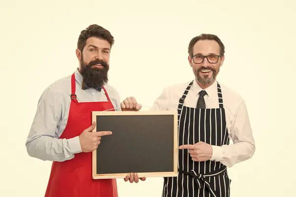 Stock image Men bearded hipster informing you. Men bearded bartender or cook in apron hold blank chalkboard. Bartender with blackboard. Hipster bartender show blackboard copy space. Bar cafe staff. Hiring staff.