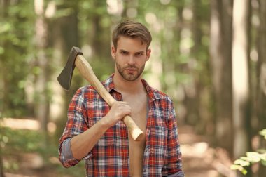 Unshaven guy in lumberjack shirt holding axe on shoulder forest background.