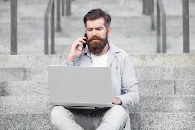 Kafası karışık adamın dışarıda online video iletişimi var. Adamın dışarıda çevrimiçi video bağlantısı var. Adamın merdivenlerde online video bağlantısı var. Adamın dizüstü bilgisayarında video bağlantısı var..