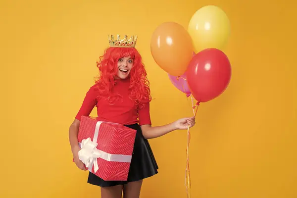 stock image glad girl in crown with present box and party balloon on yellow background.