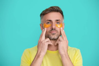 Göz altı kırışıklık önleyici krem sürmüş olgun bir adam. Yüz güzelliği cilt bakımı. Kırışıklık önleyici göz bandı olan adam mavi üzerine izole edilmiş. Yenilenme etkisi. Kozmetoloji. Cilt bakımı.