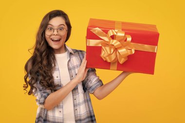 Şaşkın ergen çocuk hediye kutusunu tutuyor. Sarı arka planda izole edilmiş genç bir çocuk. Gözlüklü genç bir çocuk. Stüdyodaki gençlik fotoğrafı..