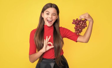Göz kırpan genç çocuk sarı arka planda bir demet üzüm tutuyor..