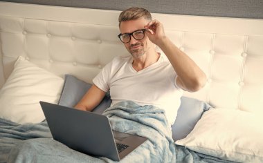 Gözlüklü olgun bir adam yatakta dizüstü bilgisayardaki e-postaları kontrol ediyor..