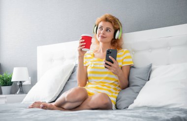 Evde akıllı telefondan müzik dinleyen mutlu kız, müzik. Dinlenen kız, yatakta çay içerken kulaklıkla müzik dinliyor..