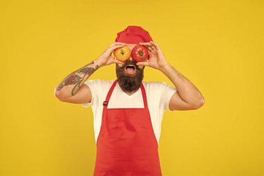 Happy guy in cooking apron and toque holding tomatoes on eyes yellow background, cook.