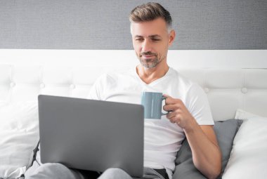 smiling mature man using computer in bed with coffee.