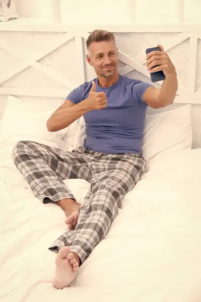 Stock image blogging man smile with phone in bedroom. blogging man with phone wear pajama. blogging man with phone in bed. photo of blogging man with phone at home.