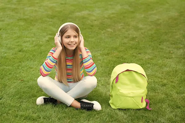 stock image Happy teen girl listening to audio course in headphones sitting on grass after school, informal education.