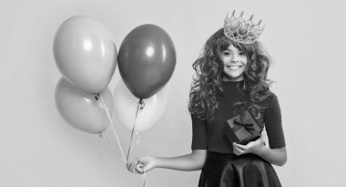 Hediye kutusuyla taç giymiş gülümseyen çocuk ve sarı arka planda parti balonu..