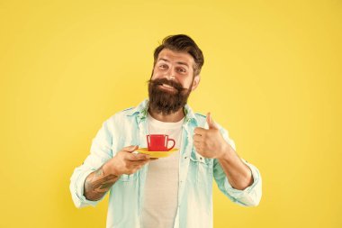 Mutlu sakallı adam sabah kahvesini içiyor sarı arka planda başparmağını gösteriyor, kahve içiyor..