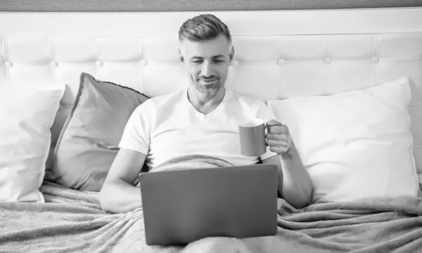 stock image happy mature man working on laptop in bed with coffee.