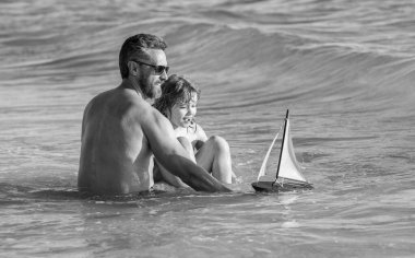 Baba ve oğul sahilde oyuncak gemiyle deniz maceraları yaratıyor. Baba ve oğul tatilde. Baba ve oğulun aile tatili. Baba ve oğulun babası açık havada birlikte oynuyorlar. Rahatlatıcı tatil.