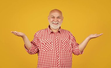 Kareli tişörtlü mutlu yaşlı adam sarı arka planda ürün sunuyor..