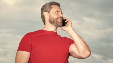 Mutlu adamın gökyüzü arka planında telefon konuşması var. Adam dışarıda telefonla konuşuyor. Adam telefonda konuşuyor, kırmızı tişört giyiyor. Bir adamın fotoğrafı telefonda konuşuluyor..