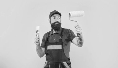 İş kıyafetleri içinde kafası karışmış sakallı adam elinde boya silindiri ve sarı arka planda fırça tutuyor..