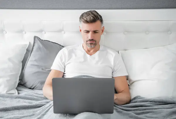 stock image concentrated mature man working on laptop in bed.