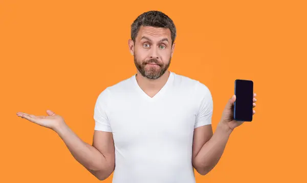 stock image confused man showing app on phone. man showing app on mobile phone with copy space. man showing phone app isolated on yellow background. man showing phone app in studio.