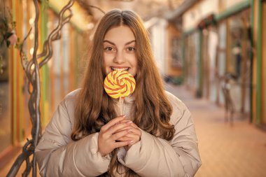 Kız dışarıda lolipop yiyor. Uzun saçlı lolipop şekerli kız. Dışarıda lolipop şekerli bir kızın fotoğrafı var. Sokakta lolipop şekerli bir kız..