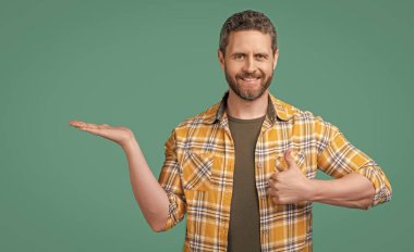 Olgun bir adam damalı gömlek giyer, ürünleri sunar ve başparmağını kaldırır. Sakallı, olgun bir adamın fotoğrafı. Olgun bir adam mavi arka planda izole edilmiş. Stüdyoda olgun bir adam.