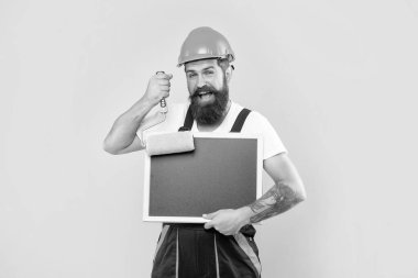 cheerful bearded man in helmet and work clothes hold paint roller and blackboard with copy space on yellow background.