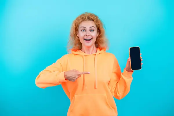 Stock image Presenting product. Advertisement of business app. Woman presenting phone screen. Young girl showing phone isolated on blue. Millennial woman. New phone app. Girl offer a product. Point finger.