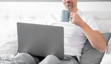 Neşeli olgun bir adam yatakta bilgisayarla kahve içiyor..