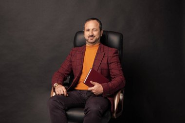 İş adamı stüdyoda planlamacıyla oturuyor. Oturan iş adamı planlamacı. İş adamının oturma planlayıcısının fotoğrafı. Ofis koltuğunda oturan iş adamı, planlayıcıyla siyah üzerine izole edilmiş