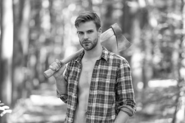 Lumbersexual guy in lumberjack shirt holding axe on shoulder forest background.